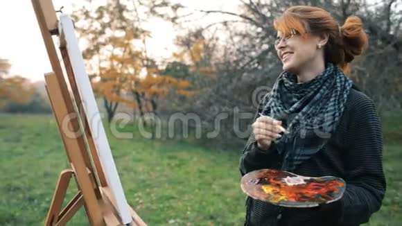 红发女孩带着一个发髻在大自然的画架上画一幅画一个年轻的女人参与了创造力享受着美丽的生活视频的预览图