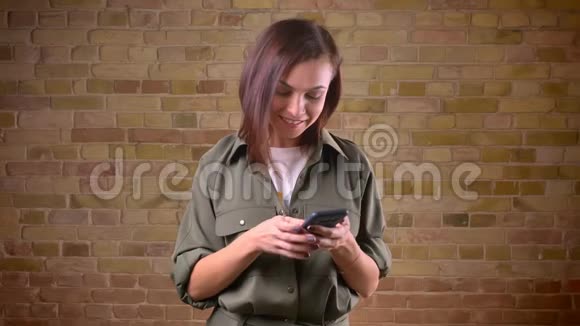 一位年轻美丽的棕色头发的女人在bricken墙背景下微笑地看着智能手机视频的预览图