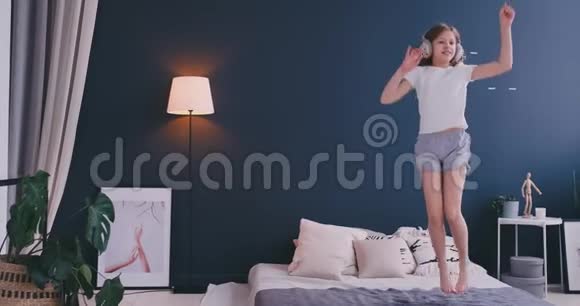 有趣可爱的小女孩戴着无线耳机跳舞梳着梳子唱歌在度假的早晨跳着床视频的预览图