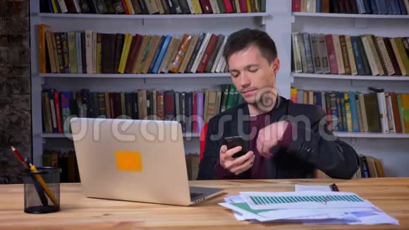 漂亮的男学生坐在笔记本电脑前在室内的图书馆里浏览电话视频的预览图