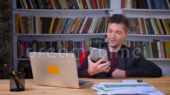 吸引人的成年快乐商人在图书馆里坐在笔记本电脑前的平板电脑上视频通话视频的预览图