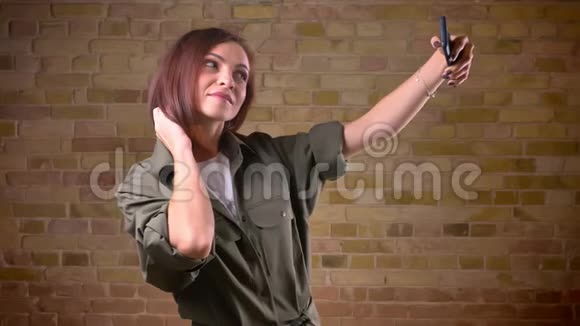 年轻漂亮的棕色头发的女人用智能手机在布里克肯墙的背景下制作自拍照片视频的预览图