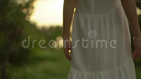 穿着白色连衣裙的苗条女孩走在夕阳的光线下沿着森林小径享受着温暖的阳光视频的预览图
