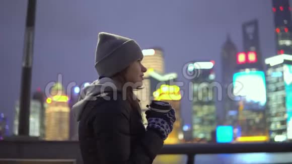 一个穿着黑色夹克和帽子的年轻女孩手里拿着咖啡在美丽的上海走来走去视频的预览图