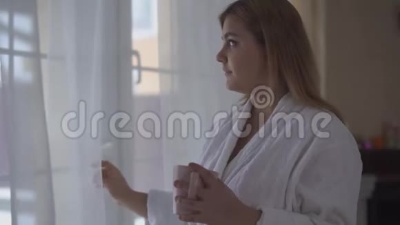 穿着白色浴袍的女孩站在大的落地到天花板的窗户附近端着一杯咖啡或茶的画像视频的预览图