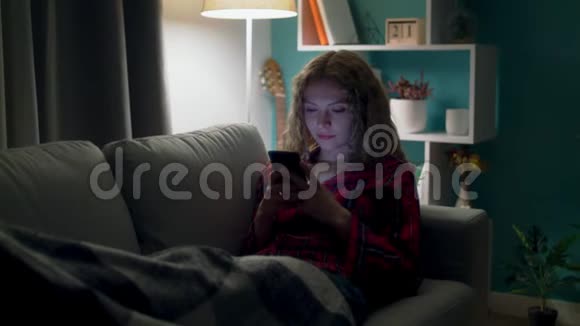 年轻女子晚上在舒适的客厅沙发上使用智能手机视频的预览图