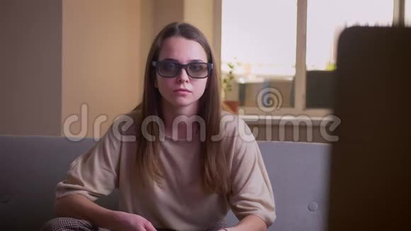 一位年轻迷人的白种人女性坐在室内沙发上看电视上的3D恐怖电影视频的预览图