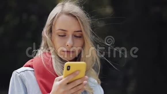 年轻美丽的女人在智能手机上使用应用程序的肖像戴着红领巾站在城市公园里视频的预览图
