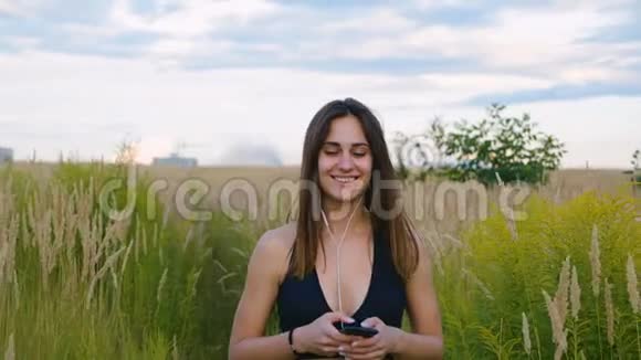 快乐的年轻女性在日落时使用智能手机和耳机在野外行走健康的生活方式女运动员使用视频的预览图