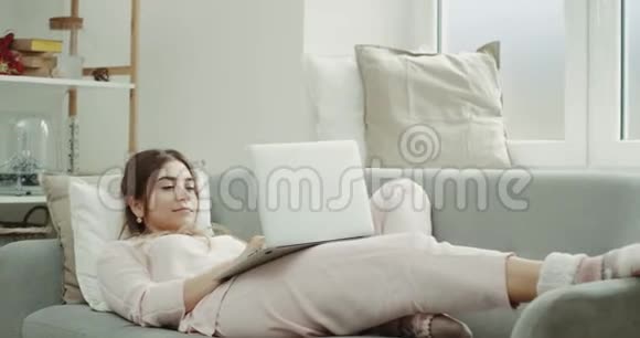 一个穿睡衣的女人在沙发上用笔记本电脑工作早上舒适的设计视频的预览图