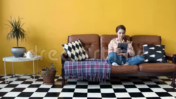 年轻漂亮的女人坐在沙发上做平板电脑库存录像设计师客厅里的年轻女子坐着视频的预览图