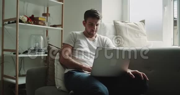 早上穿着睡衣的男人在笔记本上工作在沙发上的客厅喝咖啡现代设计视频的预览图