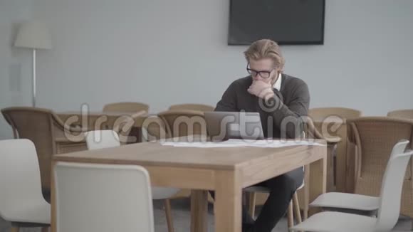 肖像画成功的金发体贴的男人戴着眼镜坐在桌子前在一间舒适的办公室前视频的预览图