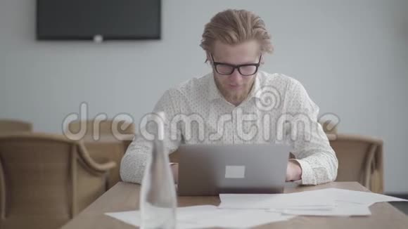 肖像英俊成功的金发体贴的男人戴着眼镜坐在桌子前在一间舒适的办公室里视频的预览图