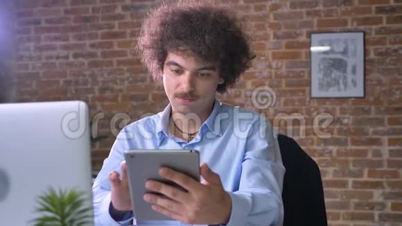 年轻开朗的书呆子商人在镜头前展示带有彩色钥匙的平板电脑微笑着坐在现代办公室里视频的预览图