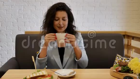 穿着蓝色夹克的肖像女孩坐在灰色沙发上吃早餐喝了一口新鲜咖啡把杯子放在桌子上视频的预览图