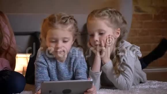 三个小白种人女孩躺在地板上在舒适的家中全神贯注地看着平板电脑的画像视频的预览图