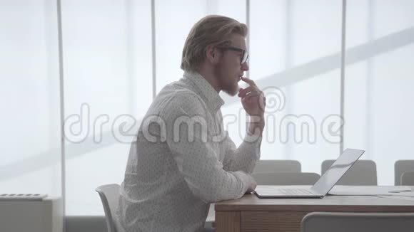一个戴眼镜的金发体贴的男人坐在桌旁坐在上网本前一间舒适的办公室里抚摸着他的眼睛视频的预览图
