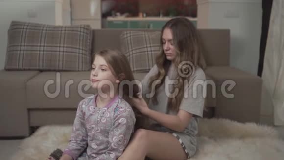 姐姐梳着小女孩的头发坐在沙发附近的蓬松地毯上姐妹关系视频的预览图