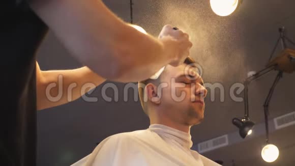 在沙龙剪发后将年轻发型师用喷雾器向男性头发喷洒水理发师梳头视频的预览图