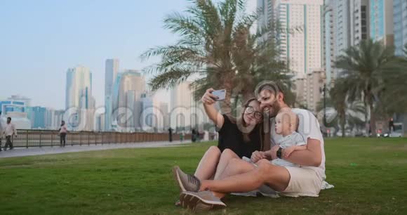 幸福的家庭两个孩子坐在公园的草地上自拍智能手机视频的预览图