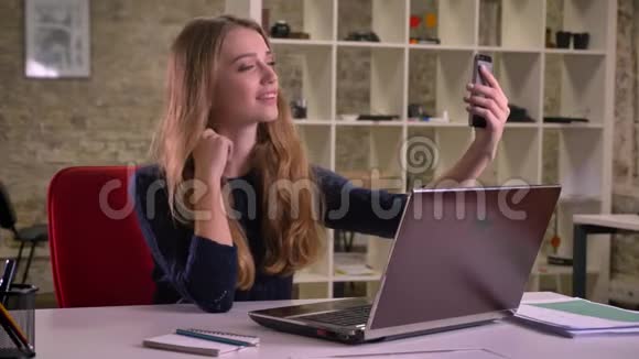 白种人金发女商人在办公室笔记本电脑前的肖像用智能手机制作自拍照片视频的预览图