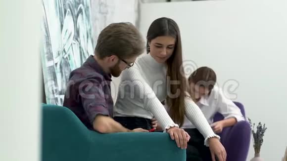 在中心的年轻人轻轻地把他的女朋友抱在扶手椅上他们看着娱乐区的电话视频的预览图