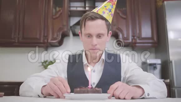 一个戴着生日帽的年轻人在厨房的一个小巧克力蛋糕上吹灭蜡烛的肖像书呆子有视频的预览图