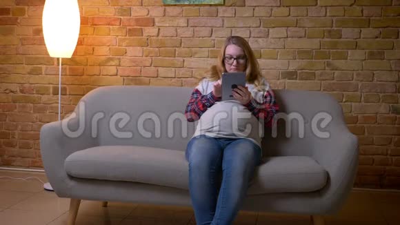 年轻怀孕女性坐在舒适的室内公寓沙发上浏览平板电脑的特写镜头视频的预览图