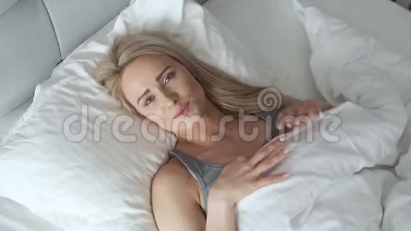 微笑的金发女人躺在床上在明亮的卧室被子下视频的预览图