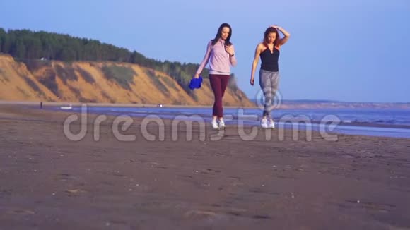 两个穿着运动服和运动鞋的女人正沿着一条大河的水边的沙滩散步视频的预览图