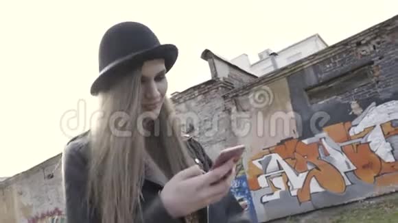 一个戴着黑帽子和皮夹克的女孩在街上靠着墙走着满是涂鸦邋遢的样子视频的预览图