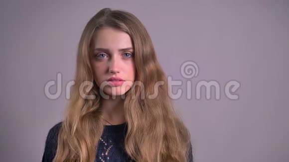 无感情的金发白种人年轻女子严肃地看着在灰色背景下眨眼进入镜头的肖像视频的预览图