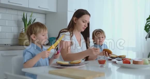 一个幸福的家庭是一个年轻漂亮的母亲穿着白色连衣裙两个儿子穿着蓝色衬衫准备着白色的厨房视频的预览图