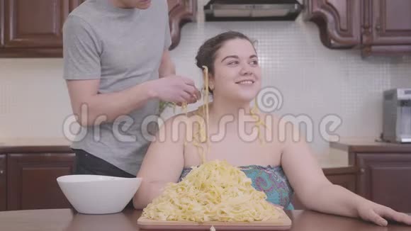 漂亮丰满的女人坐在家里的厨房里那个苗条的金发男人把面条挂在他胖女友的耳朵上视频的预览图