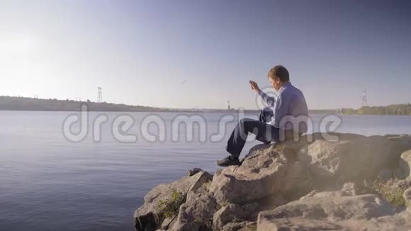 一个穿着格子衬衫的人坐在河岸上把石头扔进水里视频的预览图