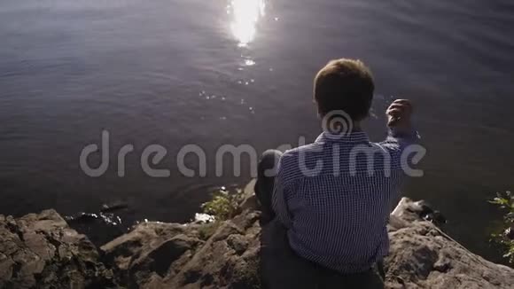 后景一个穿着格子衬衫的人坐在河岸上把石头扔进水里视频的预览图