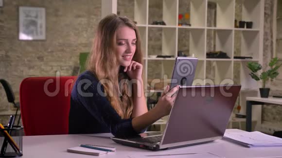 有魅力的金发白种人女商人在办公室笔记本电脑前的肖像用平板电脑制作自拍照片视频的预览图
