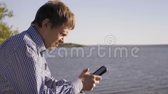 穿着格子衬衫的男人看着坐在河岸上的智能手机视频的预览图
