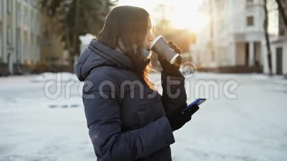 美丽的年轻女子在晴朗的冬天喝热咖啡在下雪的城市街道上使用电话视频的预览图