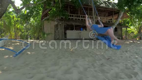 在阳光明媚的日子里穿着蓝色连衣裙的年轻女子在安静的沙滩上荡着绳子旅行概念巴拉望岛视频的预览图