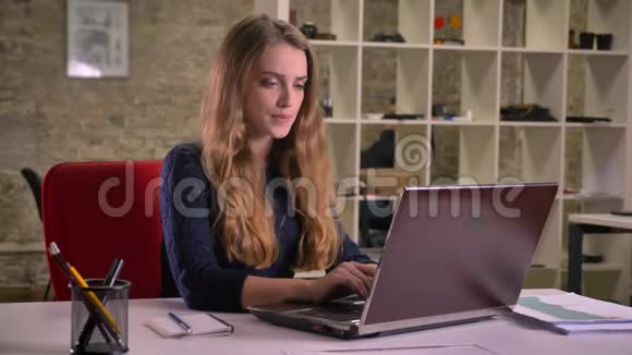 一幅白种人金发女商人的肖像她在办公室里专心地用手提电脑工作她深情地向上看着视频的预览图