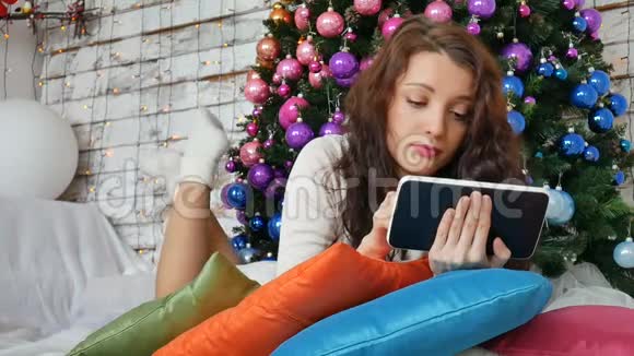 卷曲的黑发躺在圣诞树下带着平板电脑在互联网上购物新年礼物视频的预览图