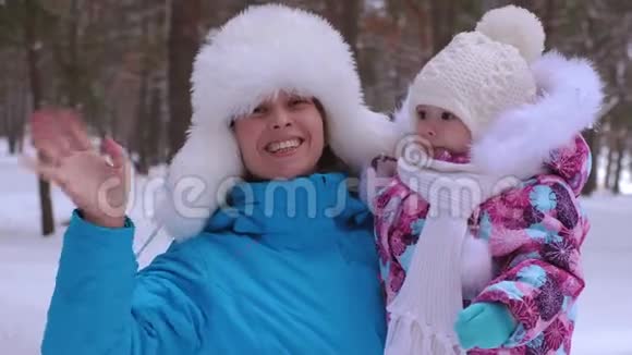 妈妈挺直了小女儿的帽子微笑着挥动她的手快乐的女人带着孩子在冬天散步笑母亲视频的预览图