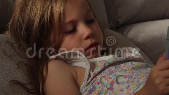 六岁的金发孩子穿着睡衣躺在沙发上拿着数码平板电脑视频的预览图