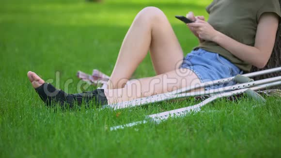 戴着拐杖的女人坐在公园里手里拿着滚动的智能手机骨折了视频的预览图