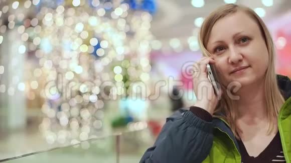 女人在智能手机上说话微笑着她正站在商场里一个灯泡不聚焦圣诞节的心情视频的预览图