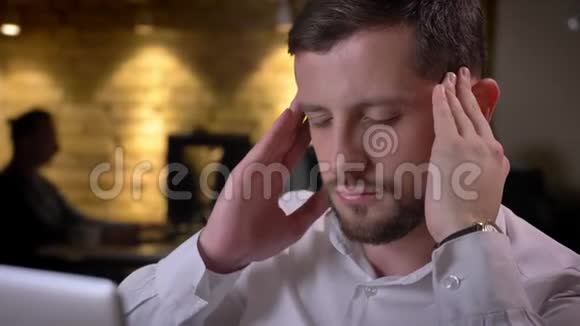 一位年轻疲惫的白种人商人在办公室的笔记本电脑前头痛和精疲力竭视频的预览图