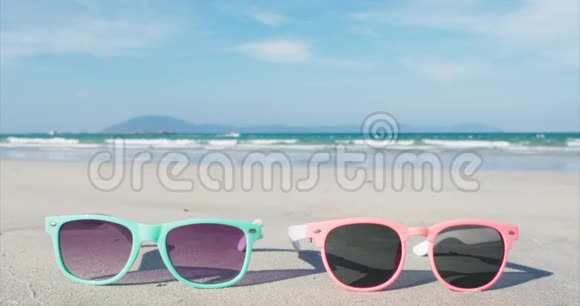 在热带海滩上粉红色和蓝色的特写太阳镜躺在白色的沙滩上靠在海洋上视频的预览图
