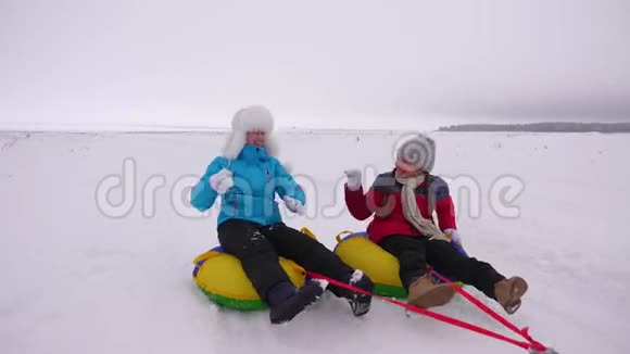 有趣的母亲和他们的女儿正沿着下雪的冬天的路骑着雪盘笑着玩雪球家庭视频的预览图